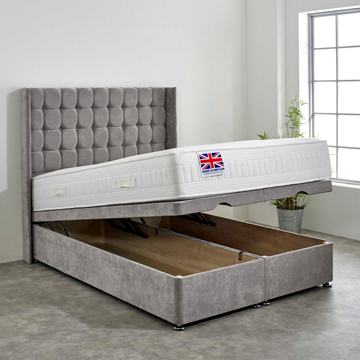 A grey upholstered cube style ottoman bed set with storage space beneath and a mattress on top, displayed in a room setting.