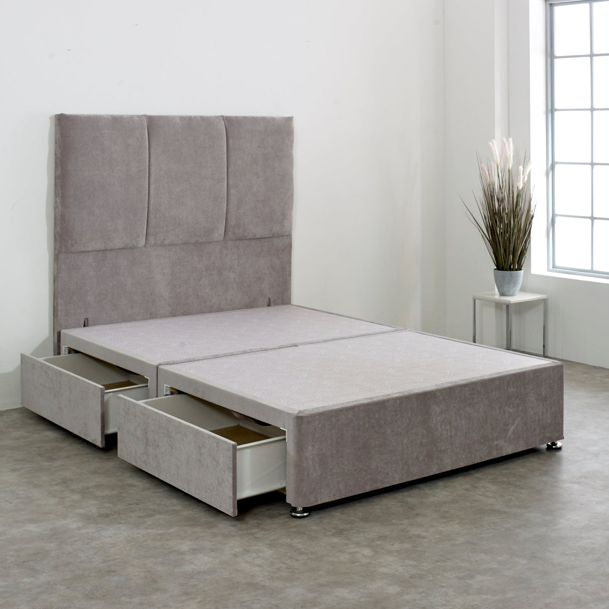 A 4ft6 divan bed base with a silver plush velvet cover, featuring two drawers at the end and chrome effect glider feet, against a minimalist room setting with a plain headboard and a plant decoration.
