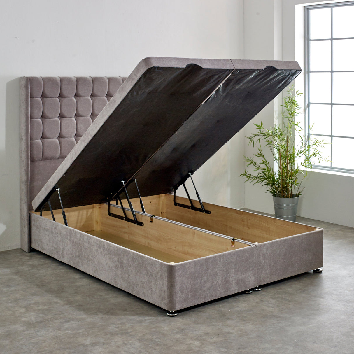 A gray upholstered ottoman divan bed base with the lid open, showing the storage compartment and the chrome effect feet.